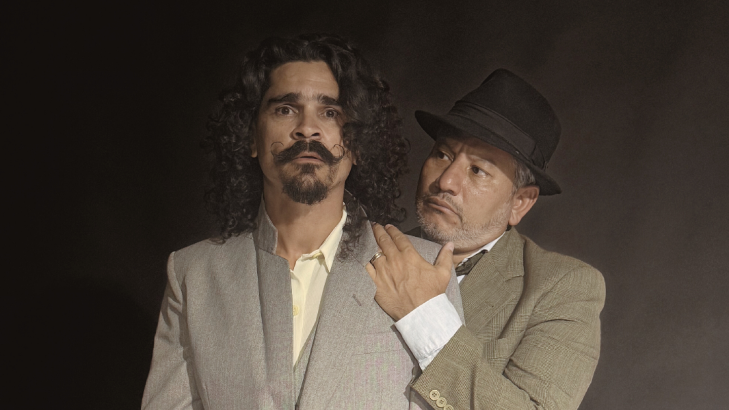 Cena teatral com dois homens caracterizados, onde um homem de chapéu coloca a mão no ombro de outro com expressão de espanto, sobre um fundo escuro