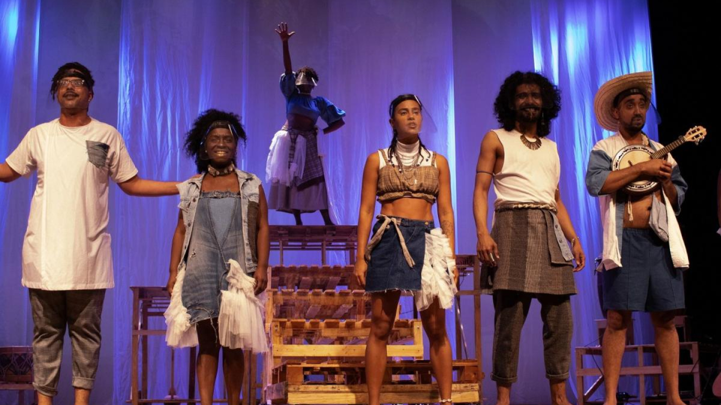 Cena de um espetáculo teatral onde um grupo de atores negros, vestidos com figurinos contemporâneos estilizados, se posiciona em frente a um cenário de tecidos azuis e estruturas de madeira