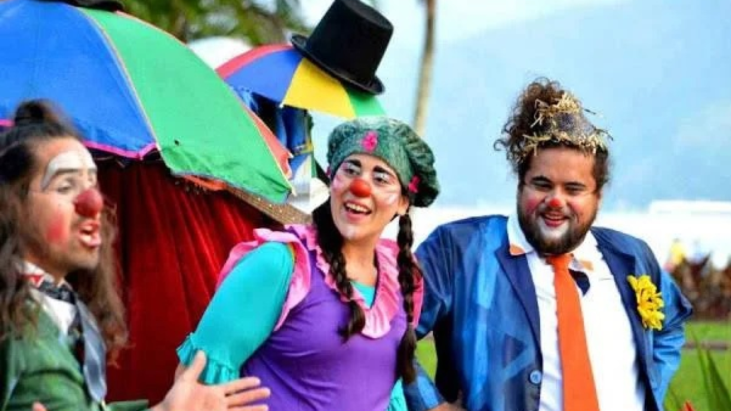 Três artistas circenses caracterizados como palhaços, com narizes vermelhos e maquiagens expressivas, realizam uma performance ao ar livre em um cenário arborizado