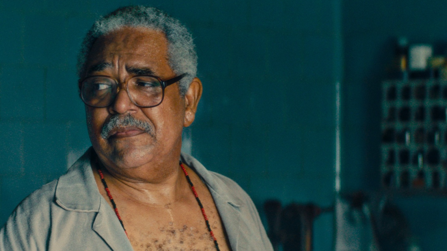 Retrato em plano fechado do ator Carlos Francisco, que aparece com expressão reflexiva e usando óculos e um colar de contas, em uma cena do filme "O Agente Secreto"