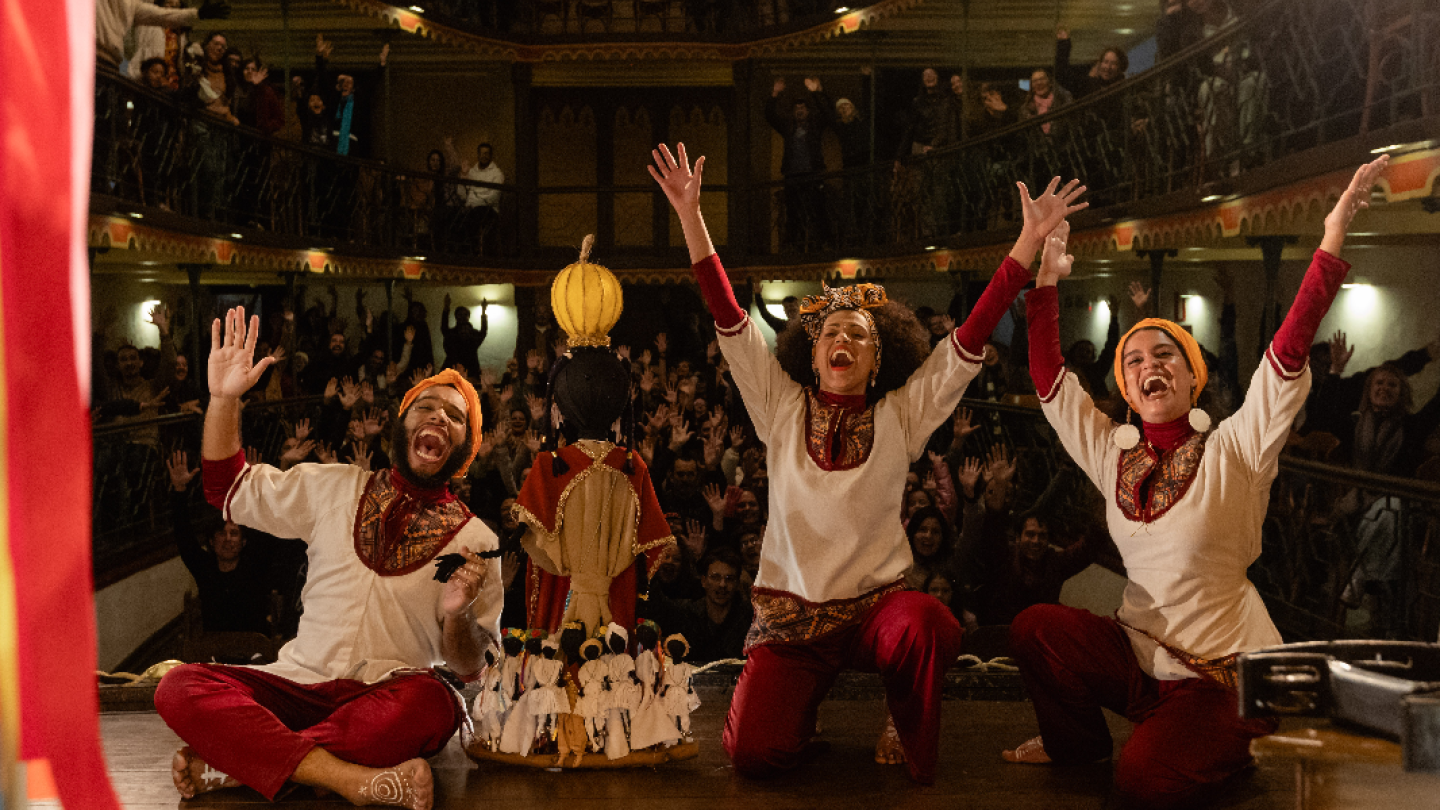 Atores com trajes tradicionais celebram com alegria e braços erguidos no palco de um teatro clássico, enquanto a plateia ao fundo retribui o gesto