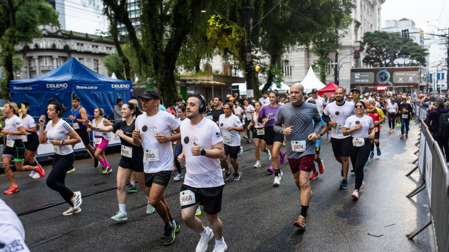 Uma multidão de corredores participa de uma prova de rua em uma via arborizada, passando por tendas de apoio e estruturas de cronometragem sob um céu claro