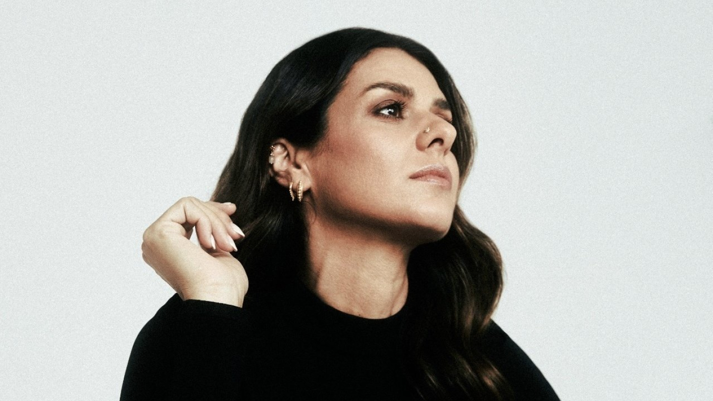 Retrato em plano médio de uma mulher de perfil, com cabelos escuros ondulados e roupas pretas, posando com a mão próxima ao rosto contra um fundo claro e minimalista