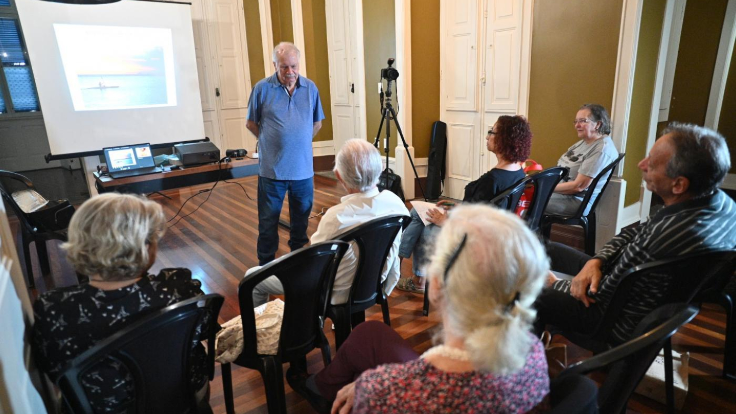 Um instrutor sênior apresenta uma aula de fotografia para um pequeno grupo de alunos em uma sala de arquitetura clássica, utilizando um projetor e uma câmera em um tripé