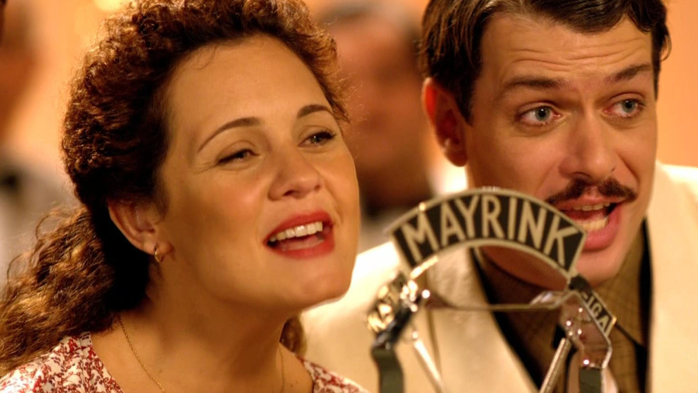 Um homem e uma mulher aparecem cantando alegremente em frente a um microfone antigo da Rádio Mayrink Veiga