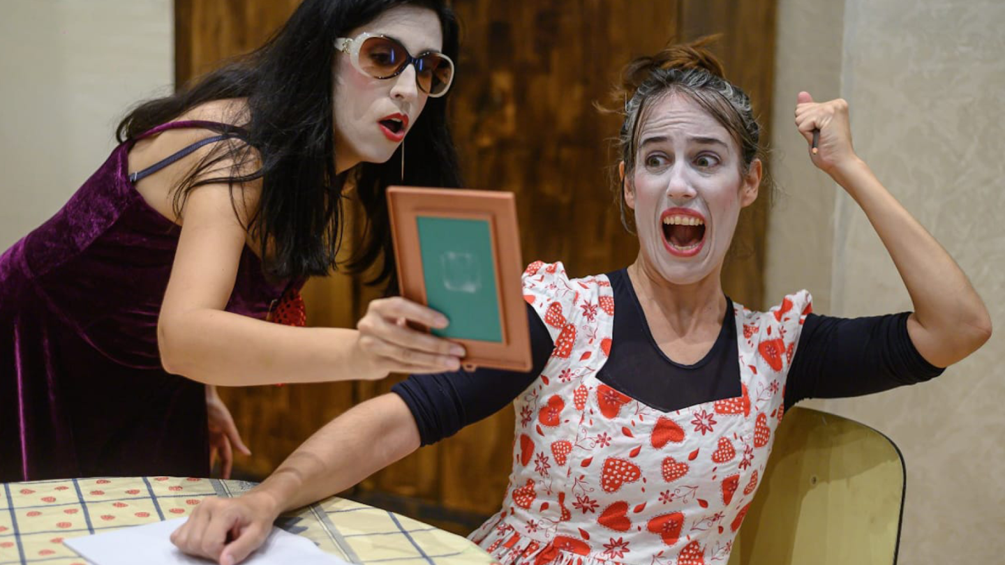 Duas atrizes com maquiagem teatral branca realizam uma cena expressiva, onde uma segura um espelho enquanto a outra reage com um grito de surpresa