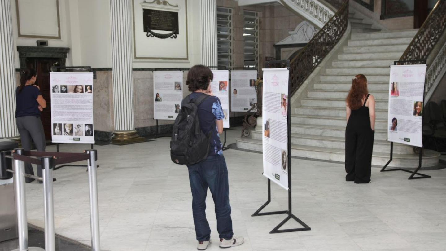 Pessoas observam uma exposição de painéis informativos no saguão de um edifício histórico, que conta com colunas clássicas e uma imponente escadaria de mármore