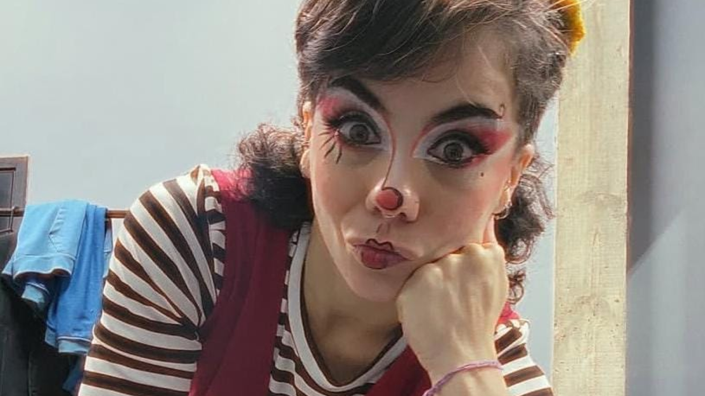 Uma artista de circo com maquiagem de palhaça expressiva, vestindo colete vermelho e blusa listrada, posa de forma lúdica com o rosto apoiado na mão