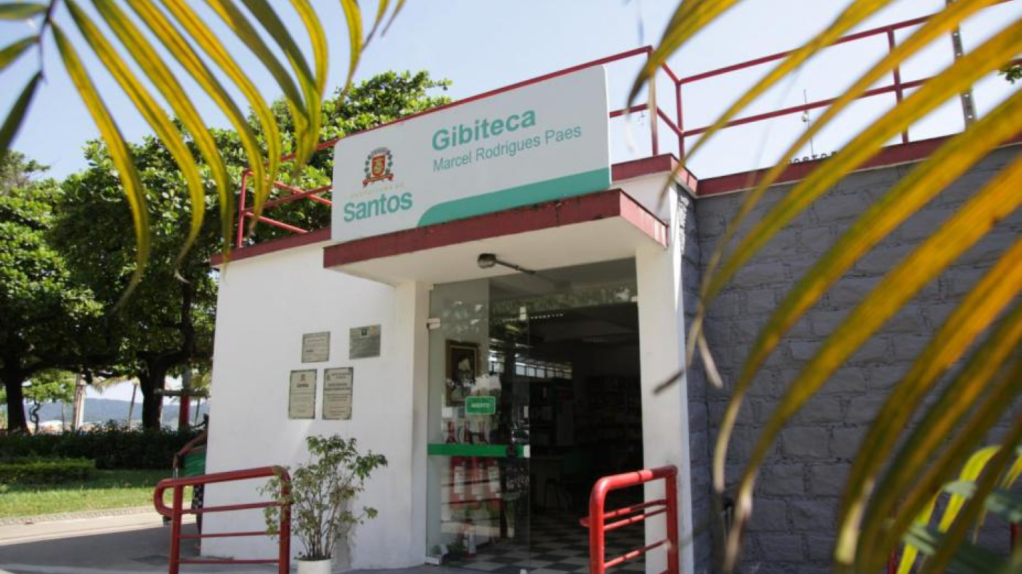 A imagem mostra a entrada da Gibiteca Marcel Rodrigues Paes, um prédio branco e cinza com detalhes em vermelho e o brasão da Prefeitura de Santos, emoldurado por folhas de palmeira em primeiro plano