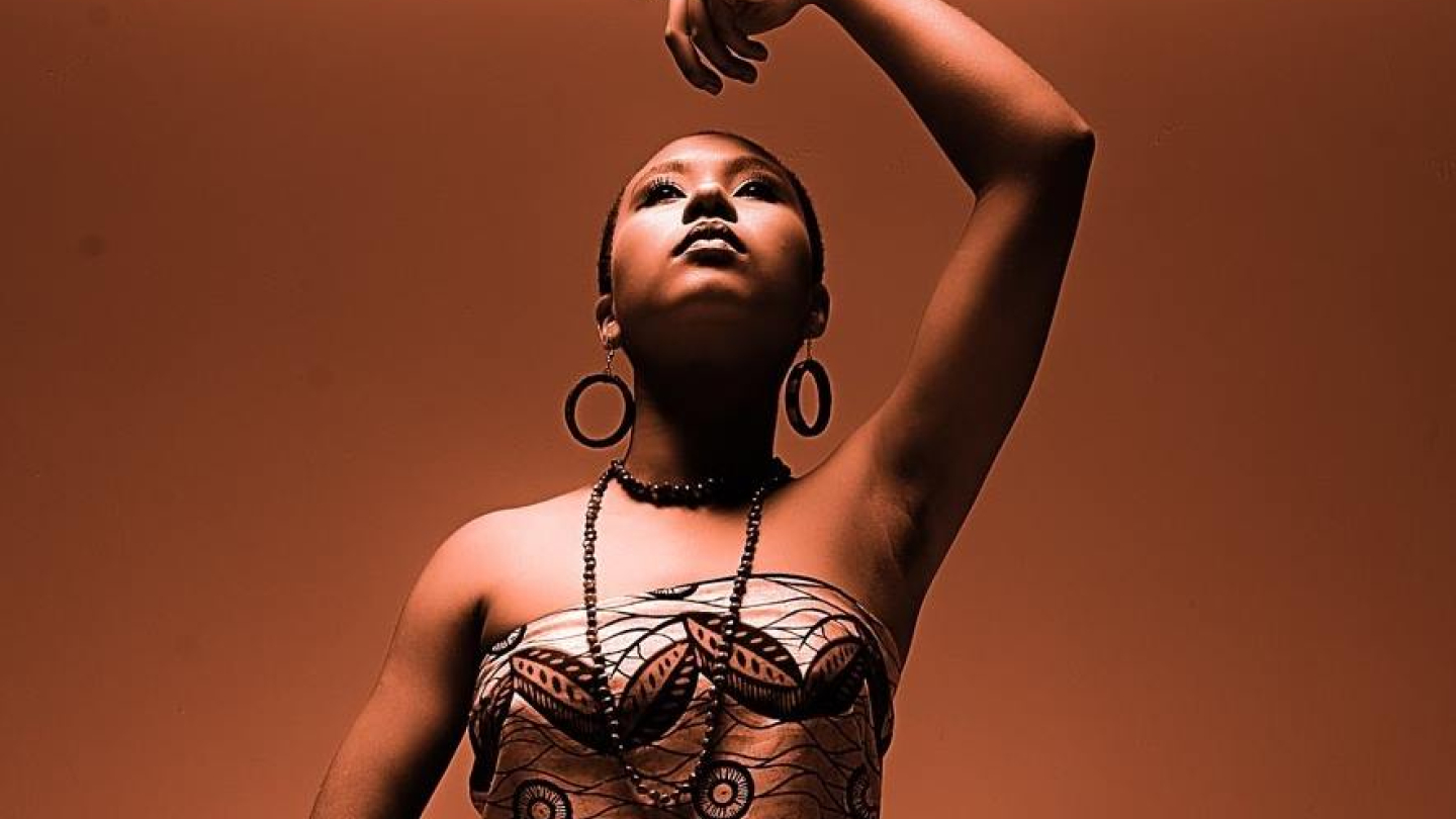 A artista Jaque da Silva posa com expressividade em um fundo em tons de cobre, vestindo uma túnica com estampas geométricas e adornos marcantes