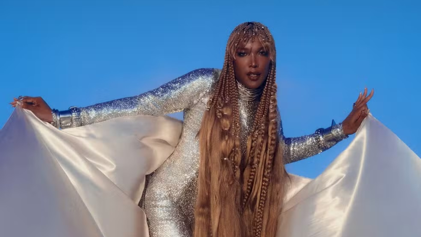 A cantora Majur posa com imponência vestindo um macacão prateado brilhante e longas tranças, destacando-se contra um fundo azul vibrante