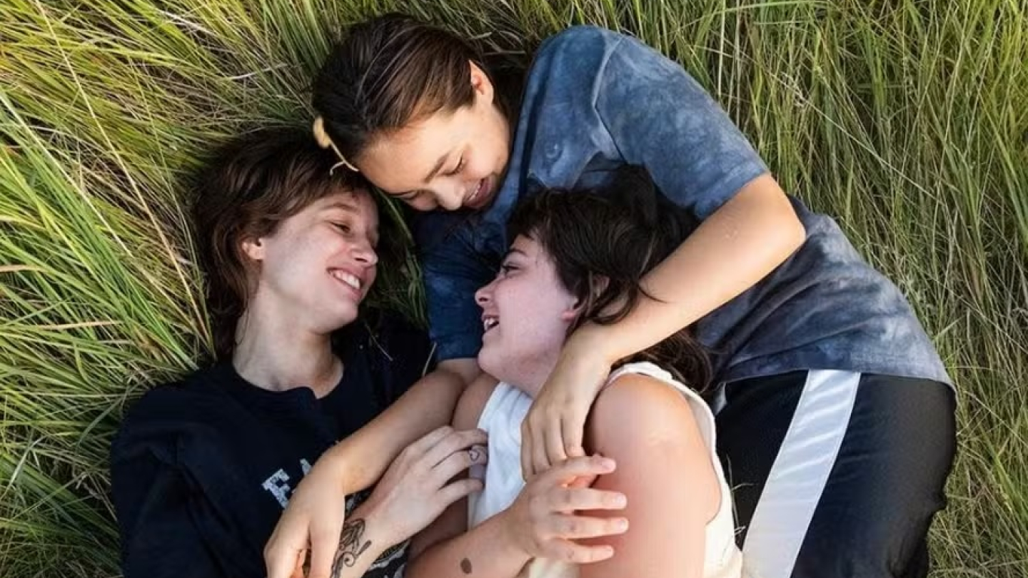 Três jovens aparecem sorridentes e abraçadas enquanto descansam deitadas sobre a grama alta