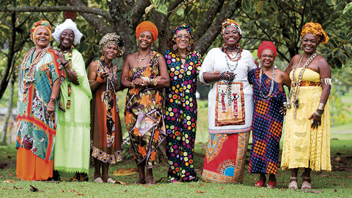 As Pastoras do Rosário posam sorridentes em meio à natureza, exibindo figurinos vibrantes, colares exuberantes e turbantes que celebram a cultura afro-brasileira