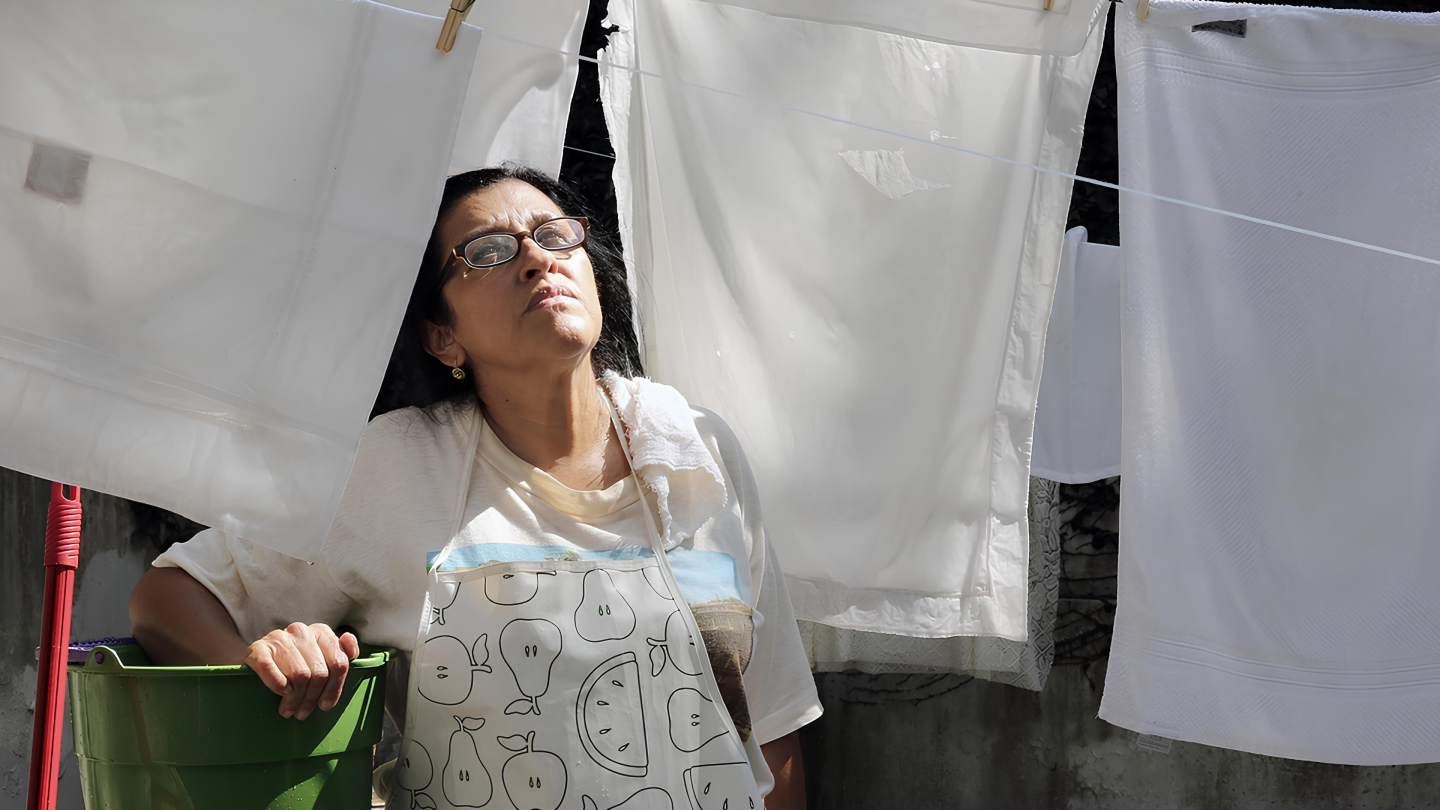 Uma mulher de óculos e avental olha para cima com expressão pensativa enquanto descansa entre lençóis brancos pendurados no varal