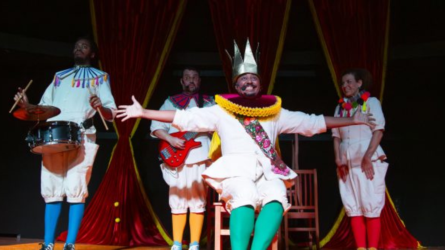 Um grupo de atores em figurinos festivos e coloridos se apresenta em um palco com cortinas vermelhas, destacando um homem de coroa dourada e braços abertos ao centro