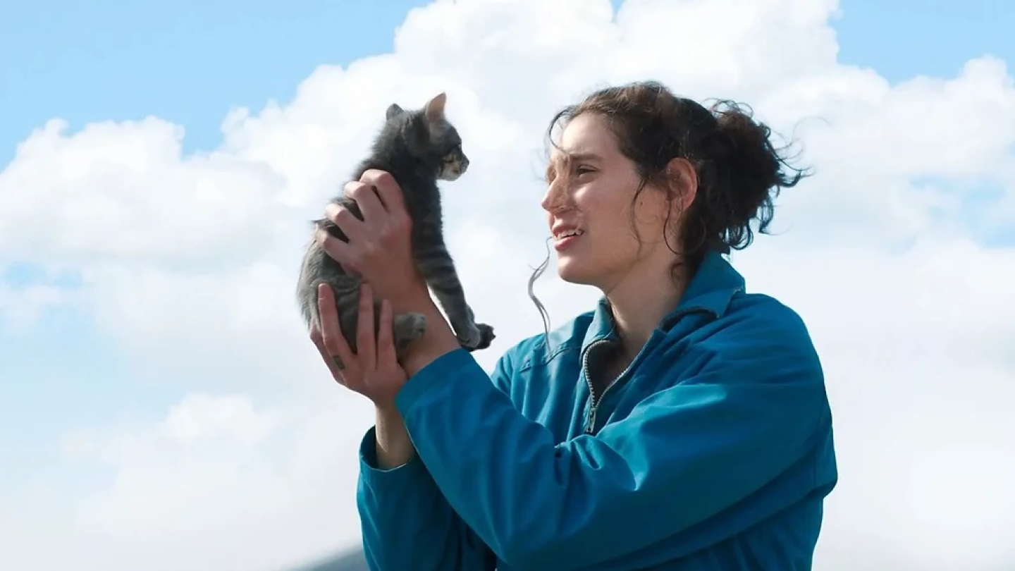 Uma mulher com expressão emocionada segura um pequeno gatinho cinza contra um céu aberto e ensolarado