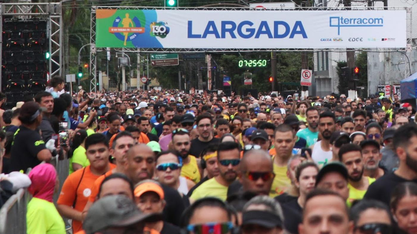 Uma multidão de corredores cruza o pórtico de largada da prova "10 KM Tribuna" sob um cronômetro digital que marca o início da competição em Santos