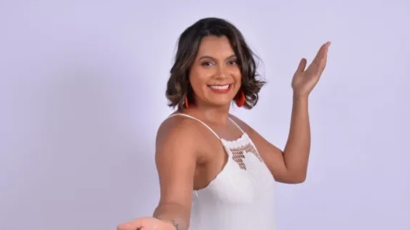 A cantora Turmalina sorri para a câmera vestindo um vestido branco, com os braços abertos em um gesto acolhedor sobre um fundo neutro