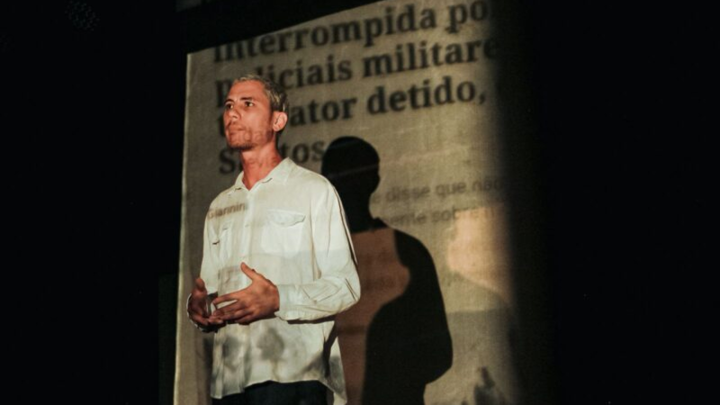 Um homem jovem com cabelo platinado e camisa branca gesticula em um palco sob a projeção de uma manchete de jornal sobre ação policial