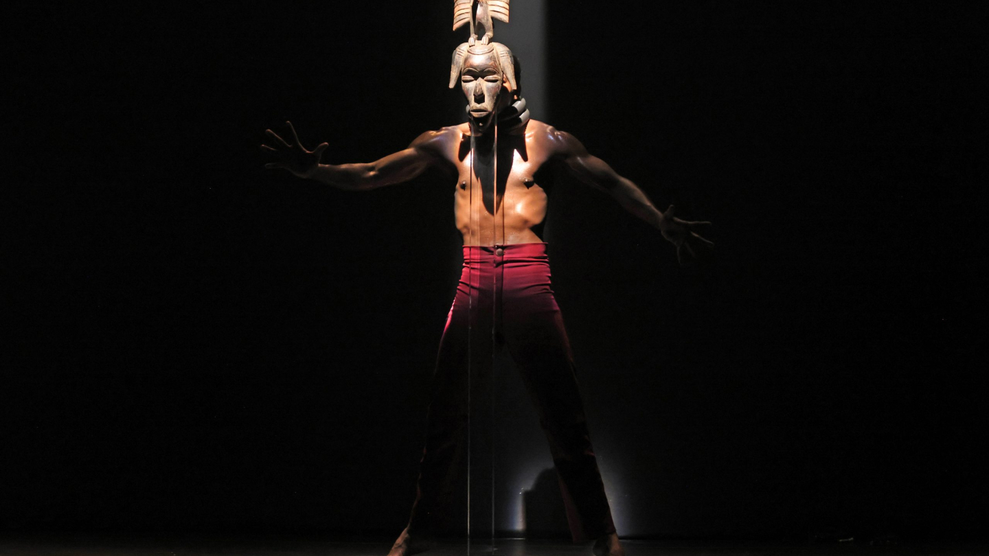 Um bailarino performa sob um feixe de luz central, vestindo calça vermelha e uma máscara tribal alongada que se estende até o chão em um cenário minimalista e escuro