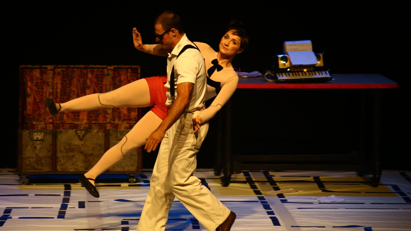 Dois atores realizam uma coreografia teatral no palco durante o espetáculo infantil "Fábrica de Brinquedos", com cenário composto por um baú e uma mesa ao fundo