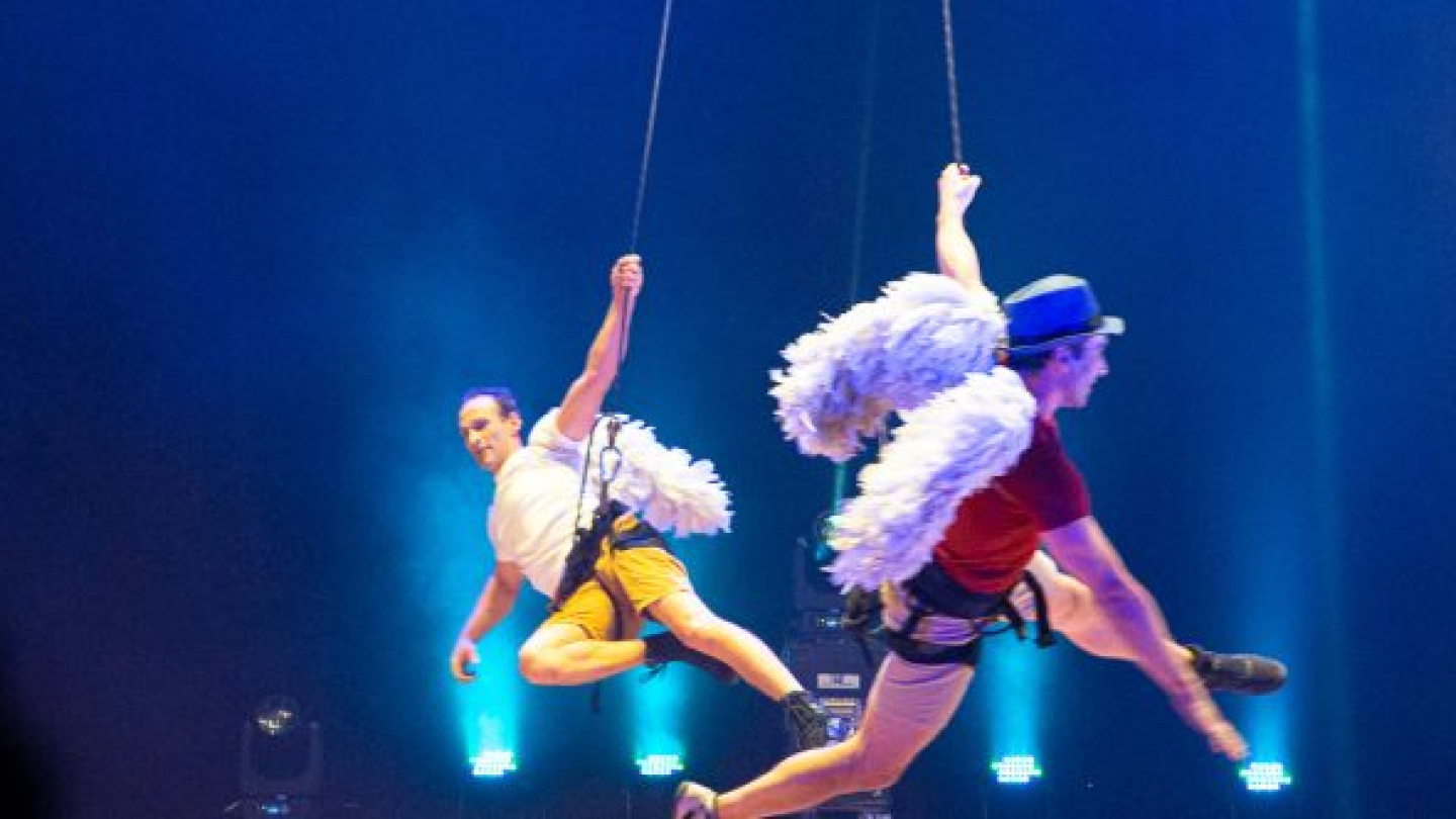Dois artistas circenses realizam uma performance aérea suspensos por cordas, vestindo grandes asas de penas brancas sob uma iluminação azulada de palco