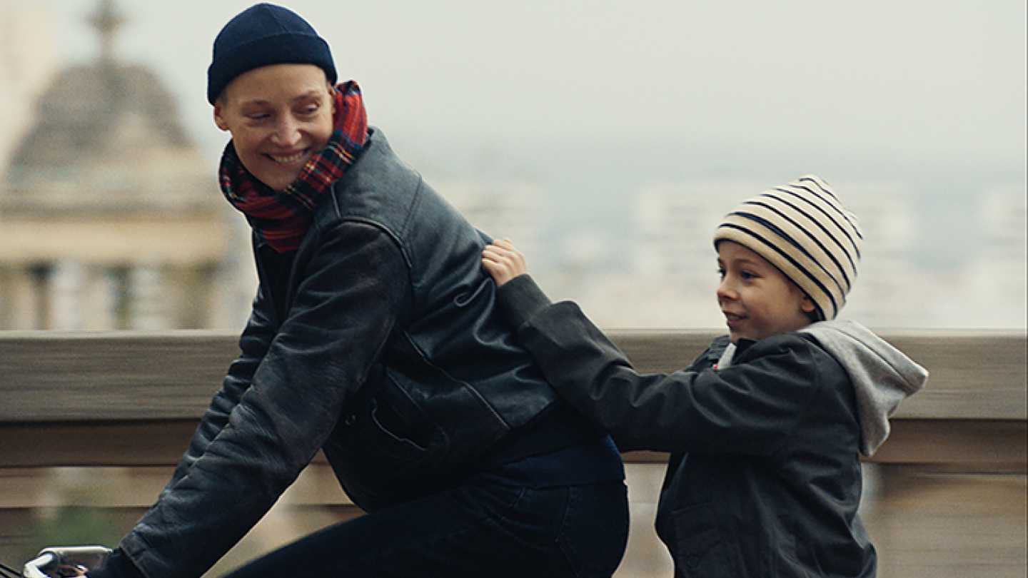 Cena do filme Me Ame com Ternura mostra uma mulher andando de bicicleta enquanto uma criança a segura pela jaqueta, em um momento afetuoso ao ar livre