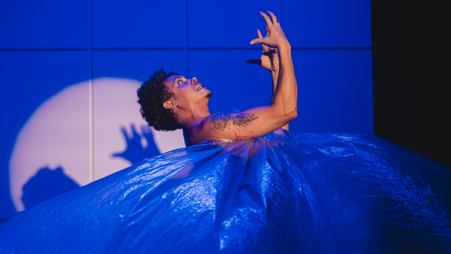 O ator Silvero Pereira realiza uma performance dramática no palco durante o espetáculo "Pequeno Monstro", emergindo de uma grande estrutura de plástico azul sob iluminação cênica