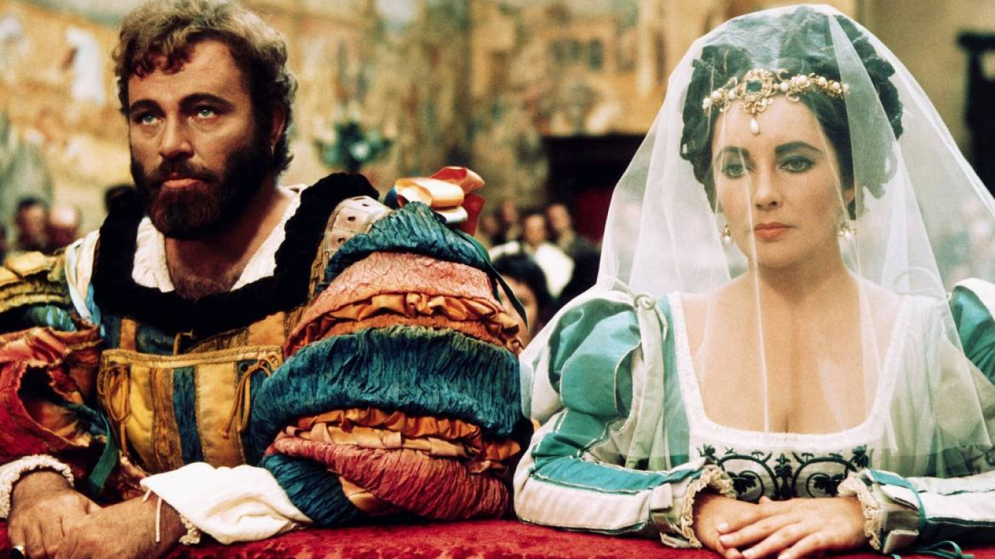 Elizabeth Taylor e Richard Burton caracterizados em trajes renascentistas para o filme "A Megera Domada", sentados lado a lado com expressões sérias durante uma cena de casamento