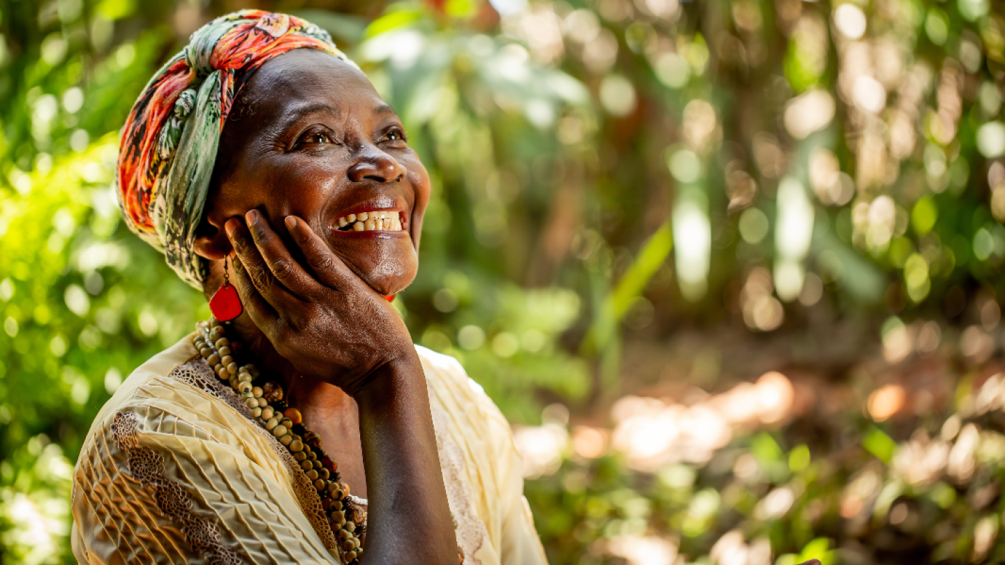 Retrato de uma mulher negra sorridente, a artista Ana Maria Carvalho, usando um turbante colorido e colares artesanais em meio a um cenário natural iluminado
