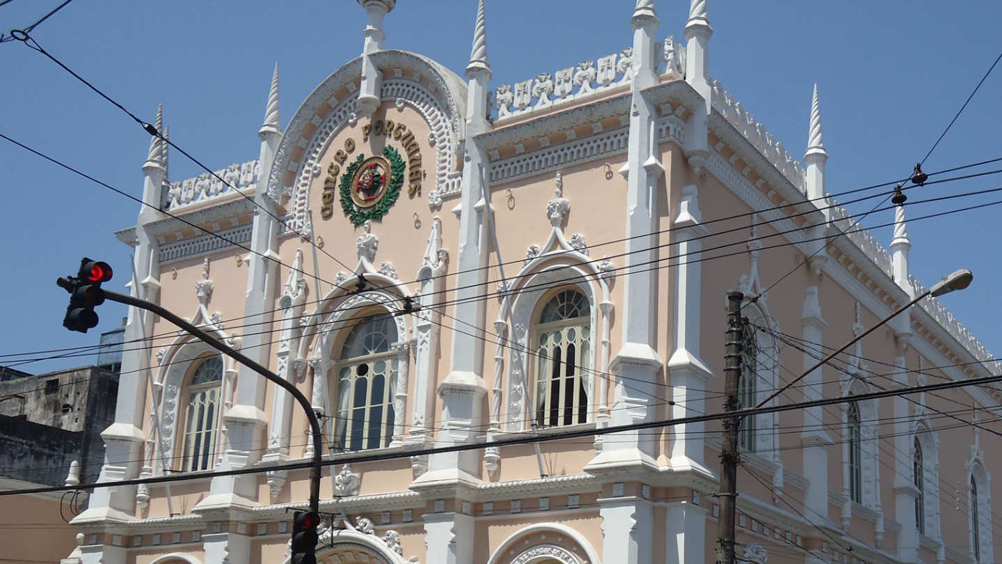 A imponente fachada de um edifício histórico em estilo neomanuelino exibe detalhes ornamentais brancos sobre paredes em tom salmão sob um céu ensolarado