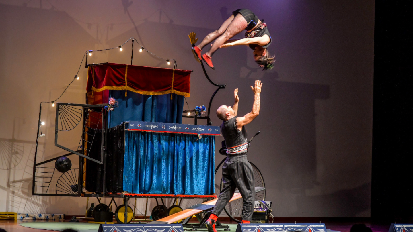 Dois acrobatas do Grupo Charivari realizam um salto em uma báscula russa em frente a um cenário circense estilizado durante uma apresentação teatral