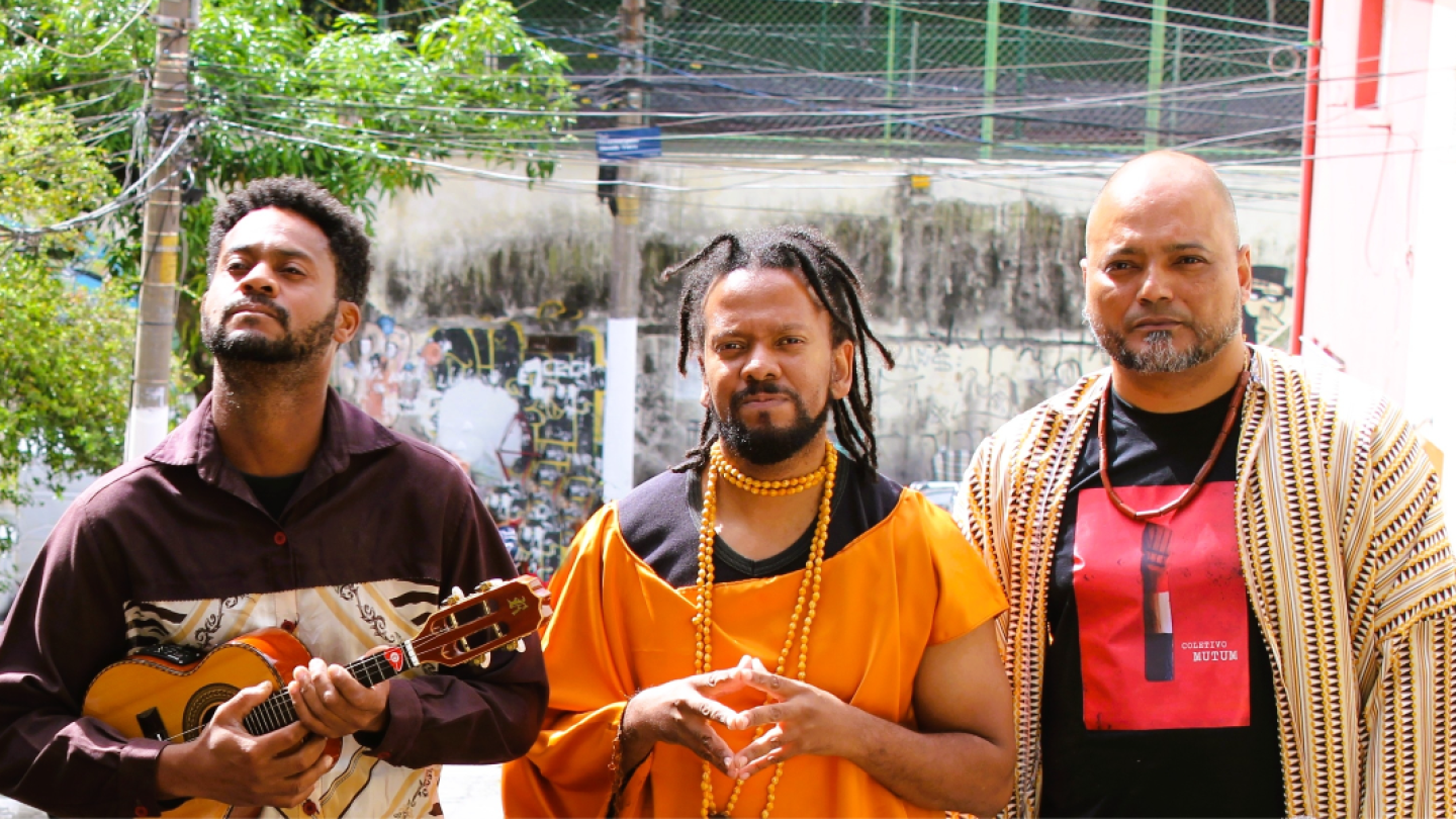 Três homens com trajes expressivos e acessórios culturais posam ao ar livre, destacando-se um deles que segura um cavaquinho