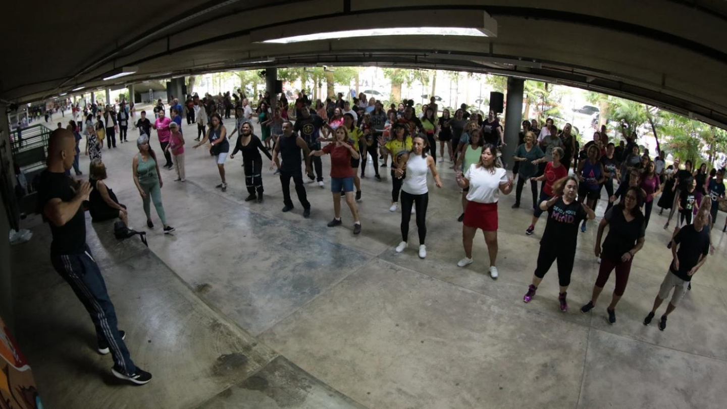 Um grande grupo de pessoas participa de uma aula de dança ou ginástica coletiva em um espaço amplo e coberto, sob a orientação de um instrutor à esquerda