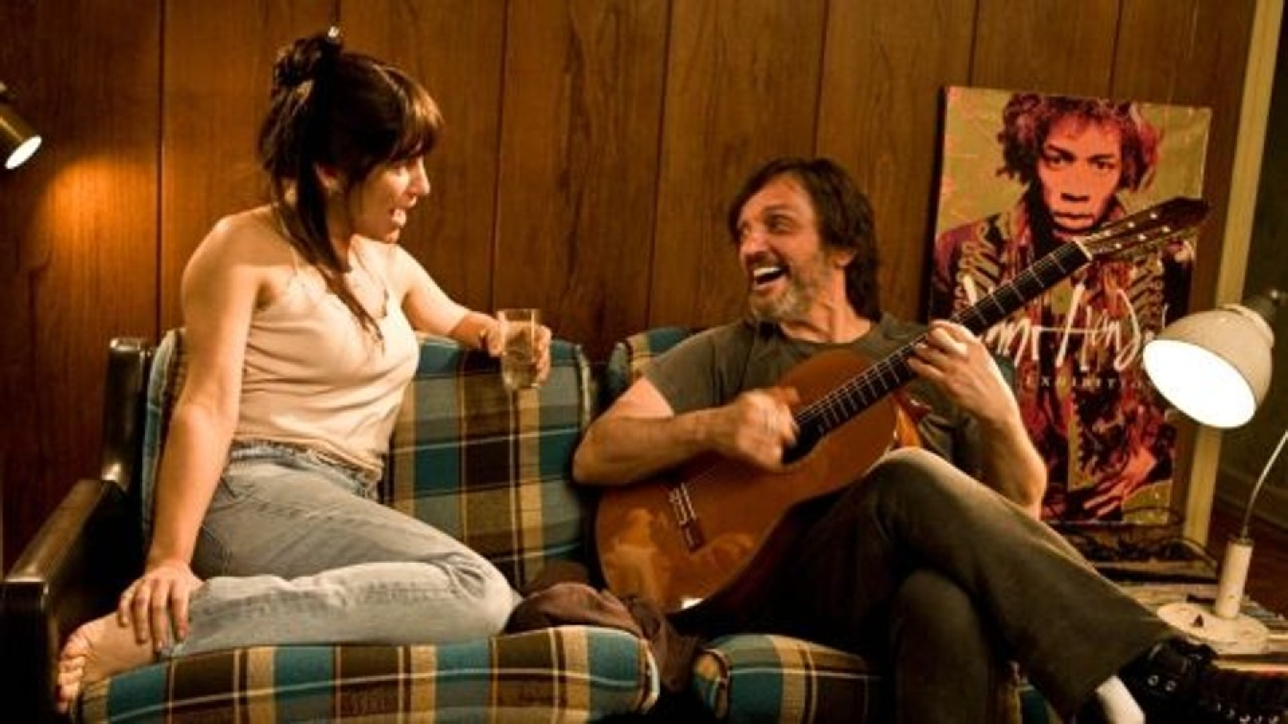 Um homem toca violão e sorri para uma mulher que o observa com um copo na mão, ambos sentados em um sofá xadrez