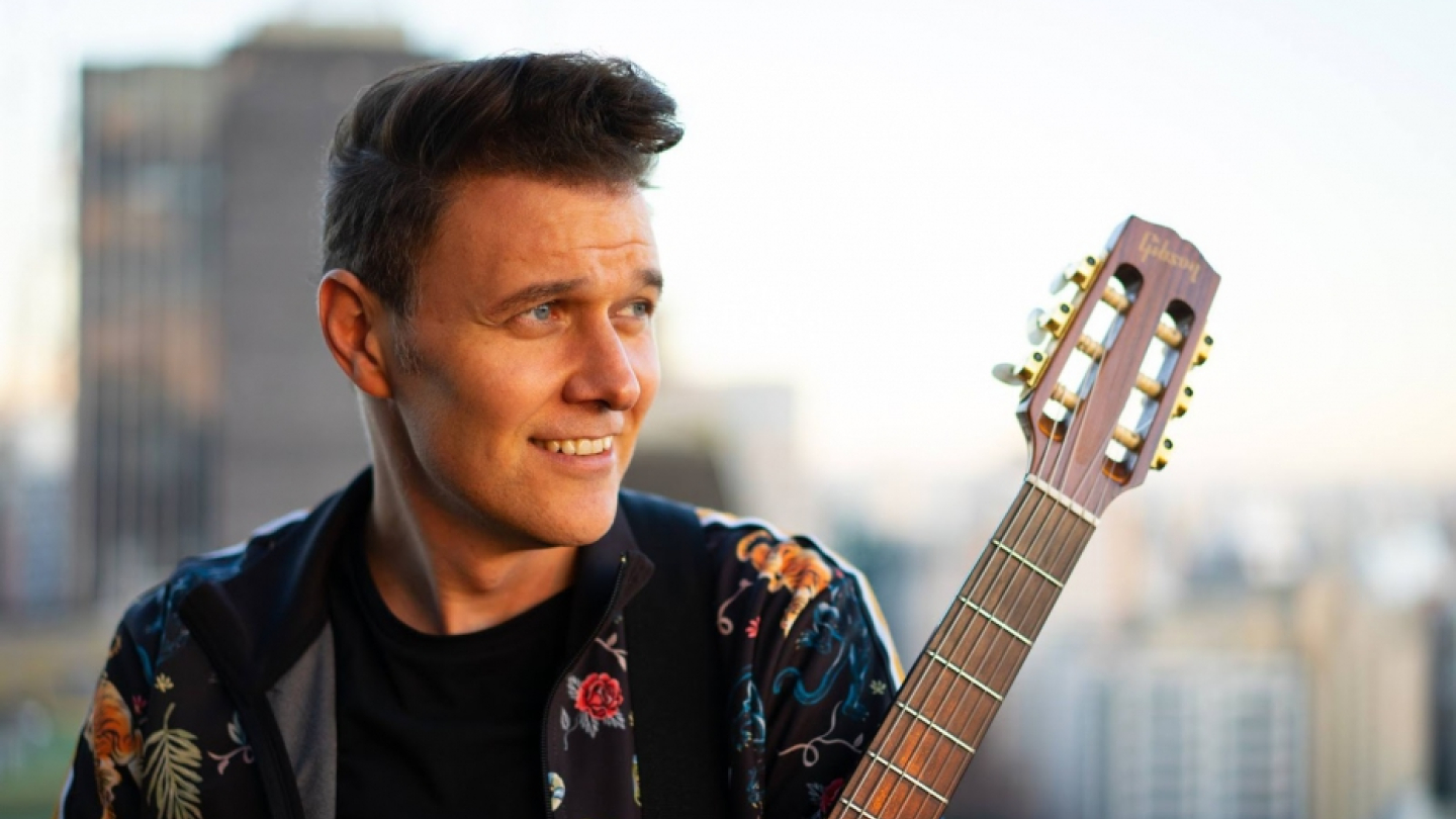O cantor e músico João Suplicy sorri enquanto segura o braço de um violão, vestindo uma jaqueta estampada com flores e tigres, tendo ao fundo uma paisagem urbana desfocada