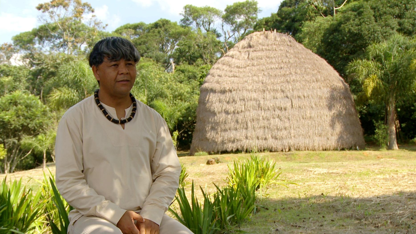 O escritor e ambientalista Kaká Werá sentado em uma área aberta com vegetação ao fundo e uma oca tradicional de palha