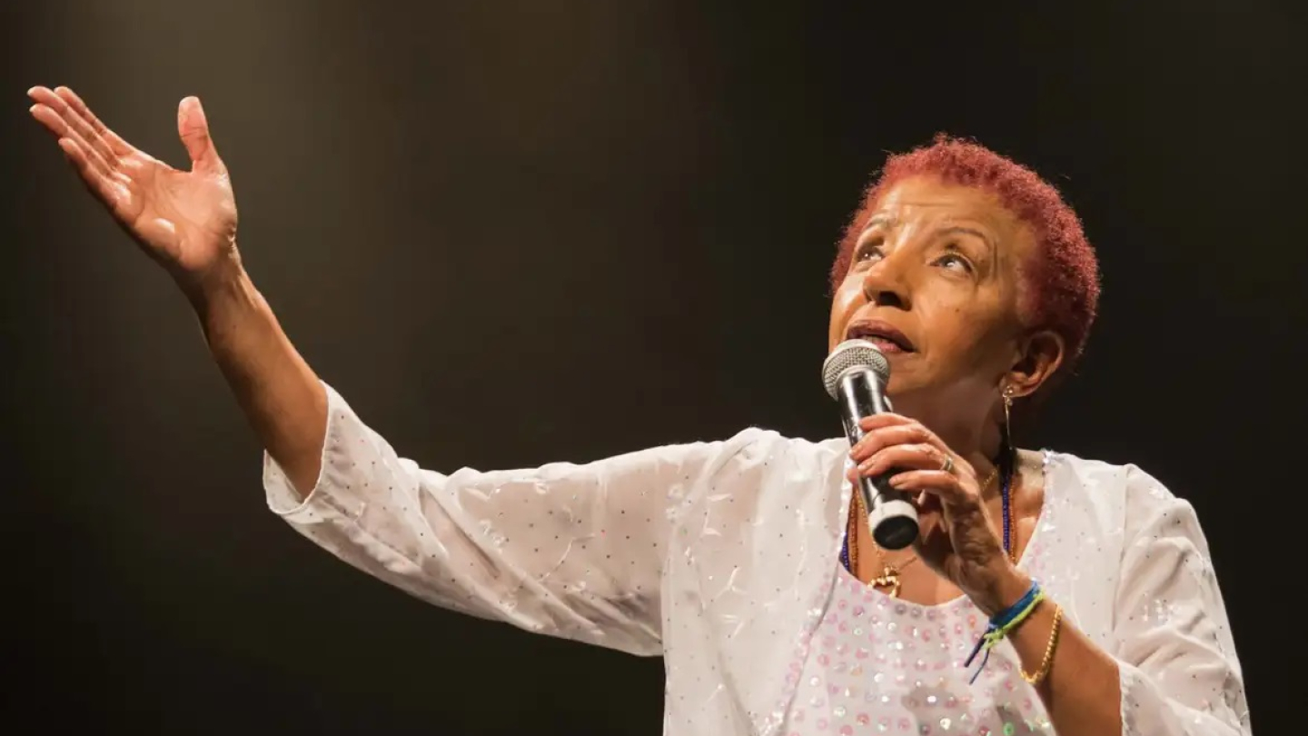 A cantora Leci Brandão se apresenta em um palco iluminado, segurando um microfone e com um braço estendido em um gesto expressivo sob o fundo escuro