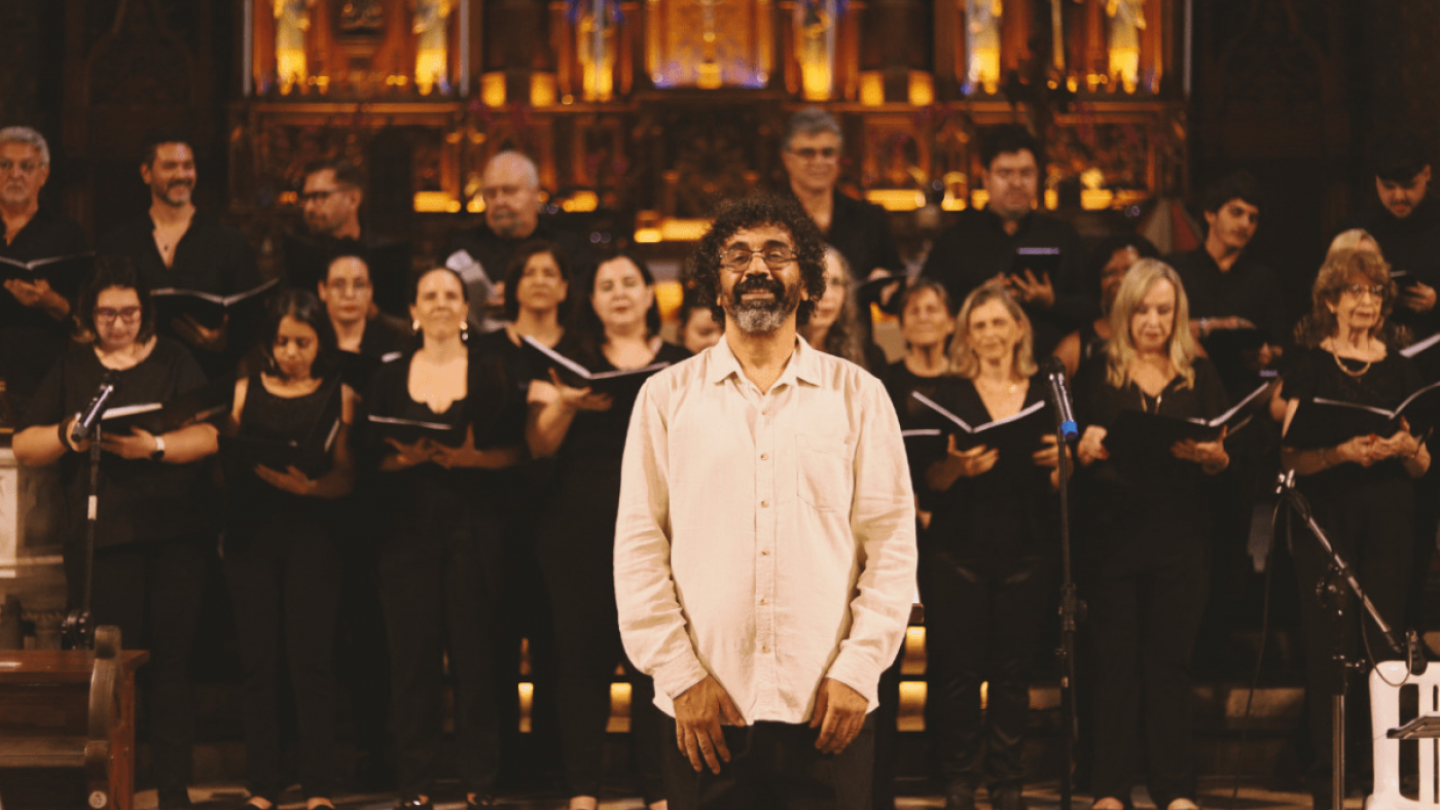 Um regente sorridente à frente de um coro vestido de preto, posicionados diante de um altar detalhado em uma igreja iluminada