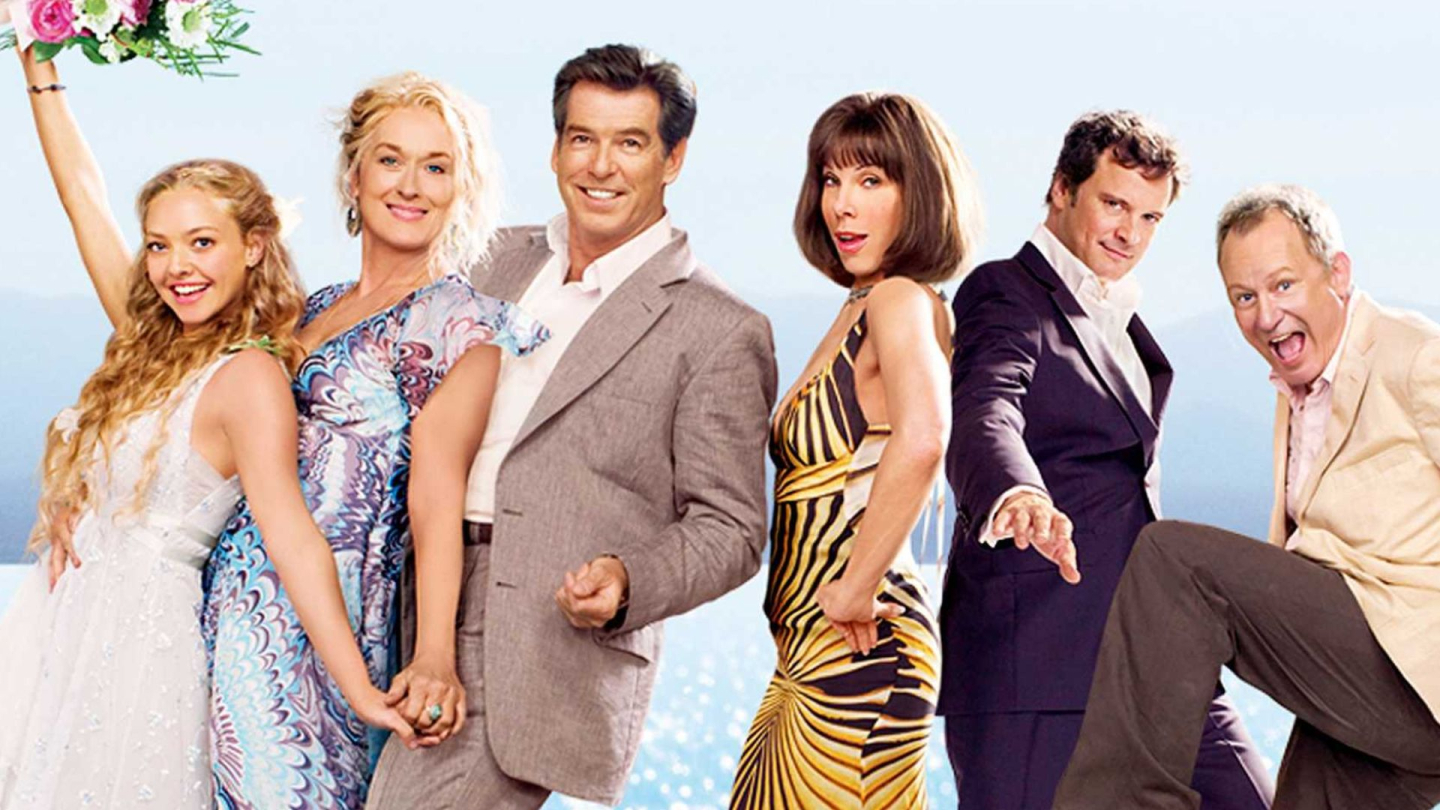 O elenco principal do filme Mamma Mia! posa alegremente em uma linha horizontal sob um céu claro, exibindo expressões entusiasmadas e trajes festivos