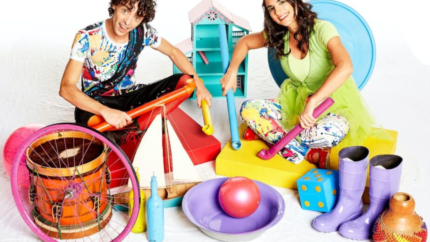 Um homem e uma mulher sorridentes posam em um cenário lúdico e colorido, cercados por diversos objetos e brinquedos como instrumentos musicais, tubos plásticos e um barco de papel