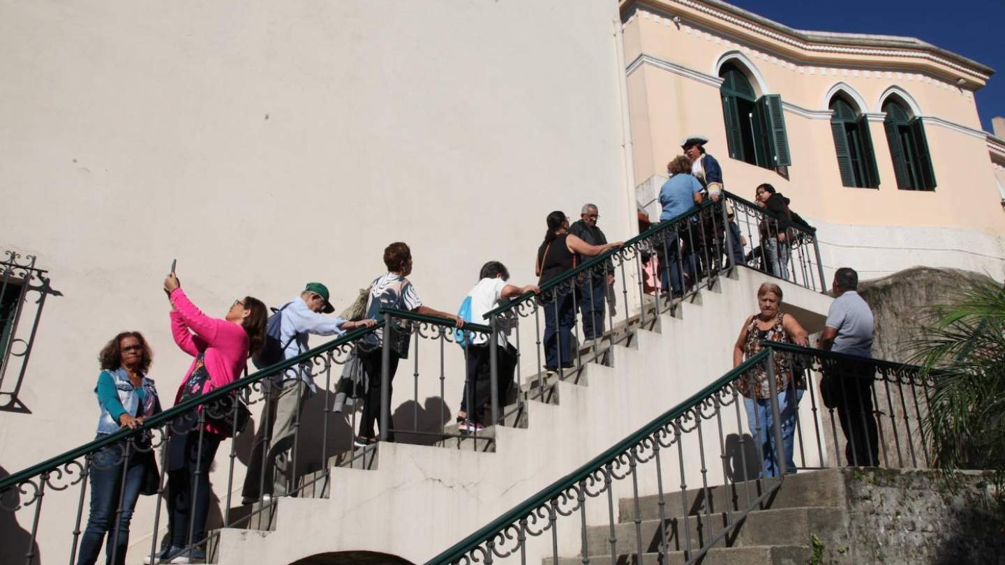 Uma fila de pessoas sobe uma escadaria externa de pedra em direção a um edifício histórico de tons claros