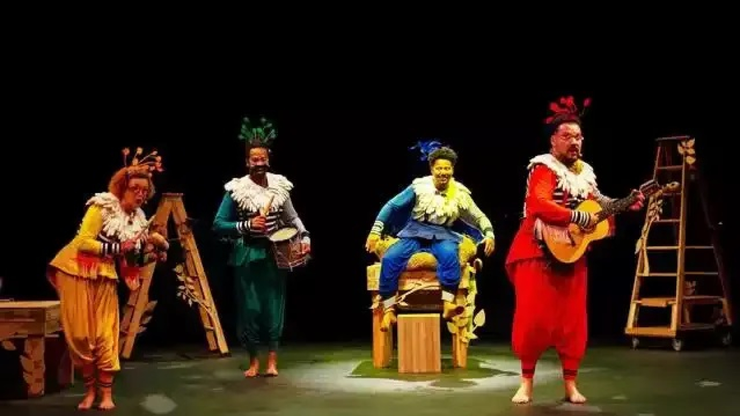 Quatro atores caracterizados com figurinos coloridos e adereços de pássaros realizam uma performance musical no palco durante a peça infantil "O Passarinho que não Sabia Voar"