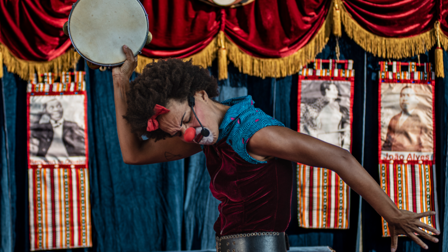Uma palhaça negra realiza uma performance expressiva segurando um pandeiro à frente de um cenário teatral com cortinas vermelhas e estandartes