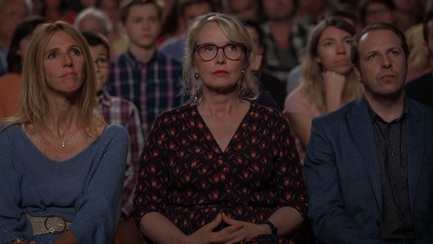 Três pessoas sentadas lado a lado em uma plateia demonstram expressões de seriedade e atenção enquanto observam algo à frente