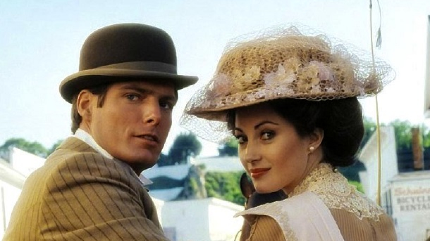 O ator Christopher Reeve e a atriz Jane Seymour olham para trás por cima dos ombros, vestindo trajes e chapéus de época no filme "Em Algum Lugar do Passado"