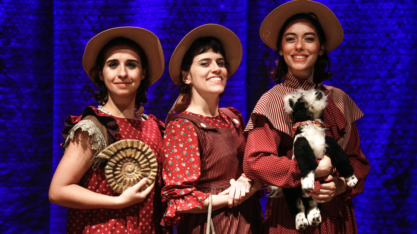 Três mulheres sorridentes, vestidas com trajes de época vermelhos e chapéus de palha, posam juntas contra um fundo azul iluminado, segurando um fóssil de amonite e um bicho de pelúcia