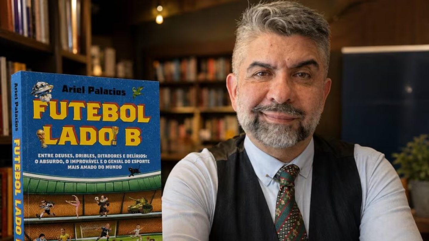 O jornalista Ariel Palacios sorri ao lado de um exemplar de seu livro, intitulado "Futebol Lado B", em um ambiente com estantes repletas de livros ao fundo