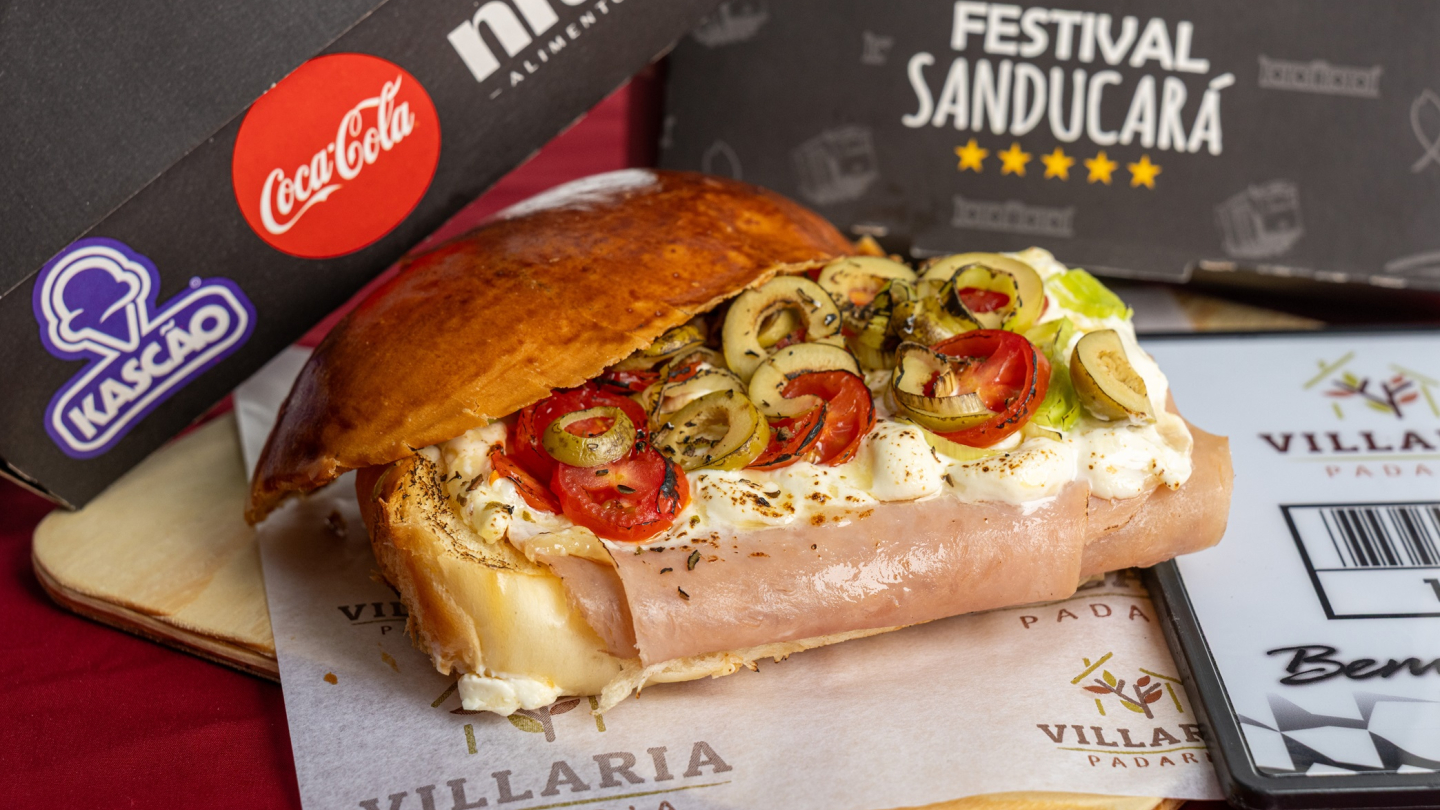Um sanduíche aberto no pão de cará recheado com presunto, queijo cremoso, tomates e azeitonas é servido sobre um papel timbrado da Padaria Villaria, acompanhado por embalagens promocionais ao fundo