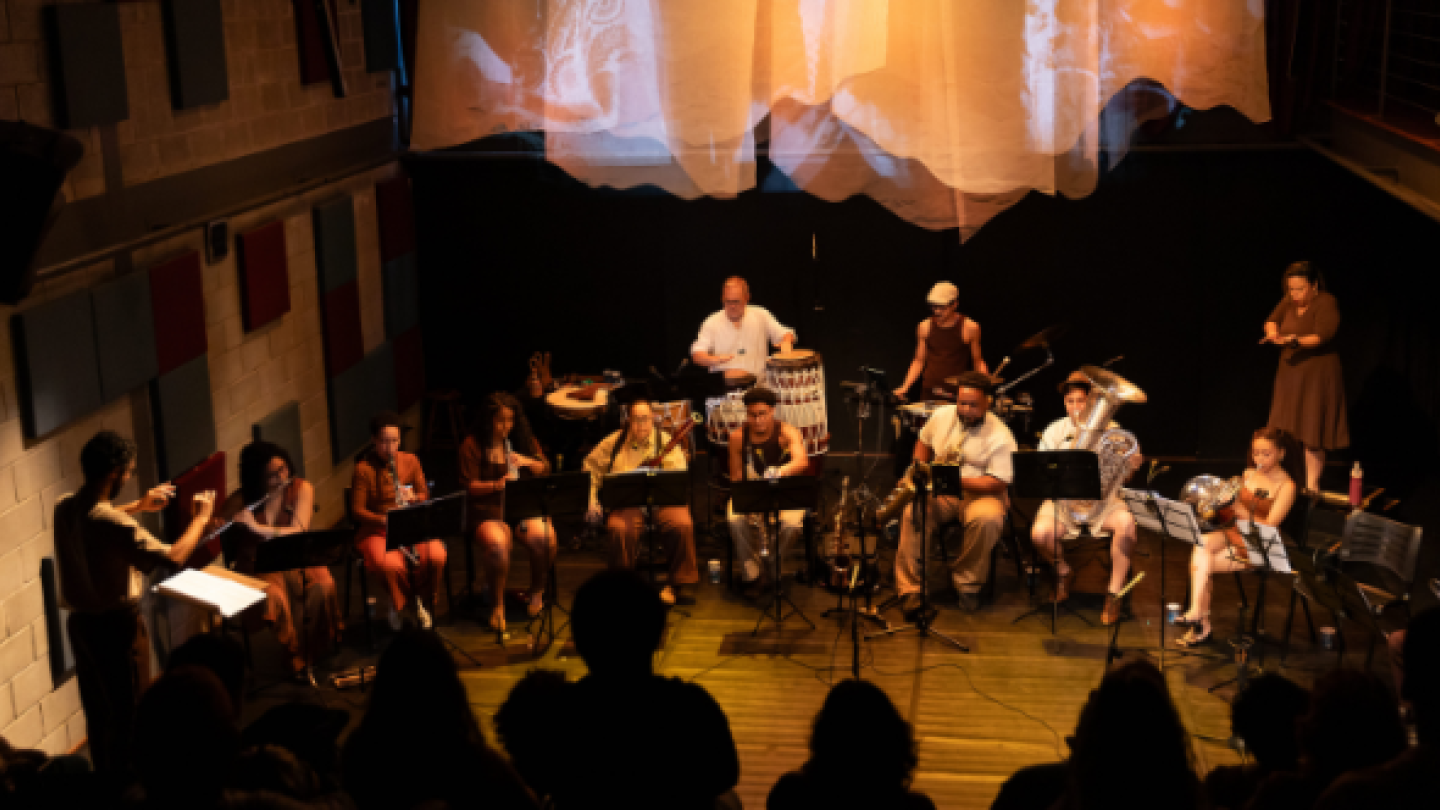 Uma orquestra de câmara se apresenta em um palco iluminado por tons quentes diante de uma plateia em silhueta, sob uma projeção de tecido abstrata suspensa ao fundo